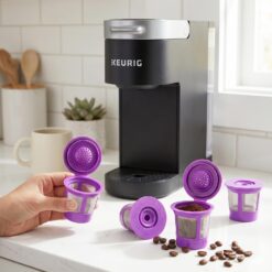 Four purple reusable Keurig coffee filter pods with coffee grounds and beans beside a Keurig brewer.