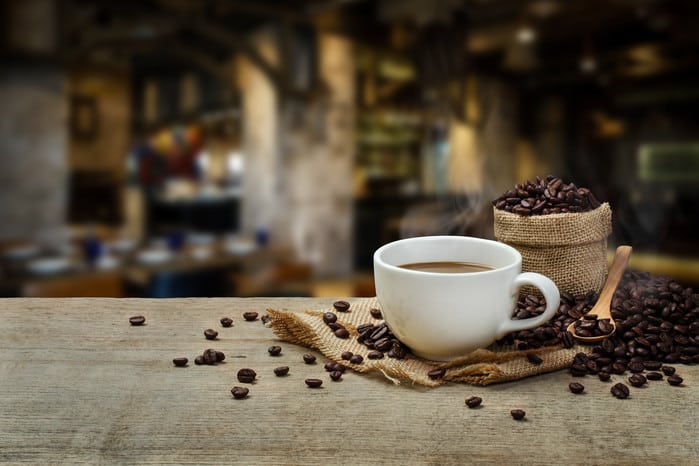 Coffee cup, beans, and burlap sack on a wooden table in a blurred cafe setting.