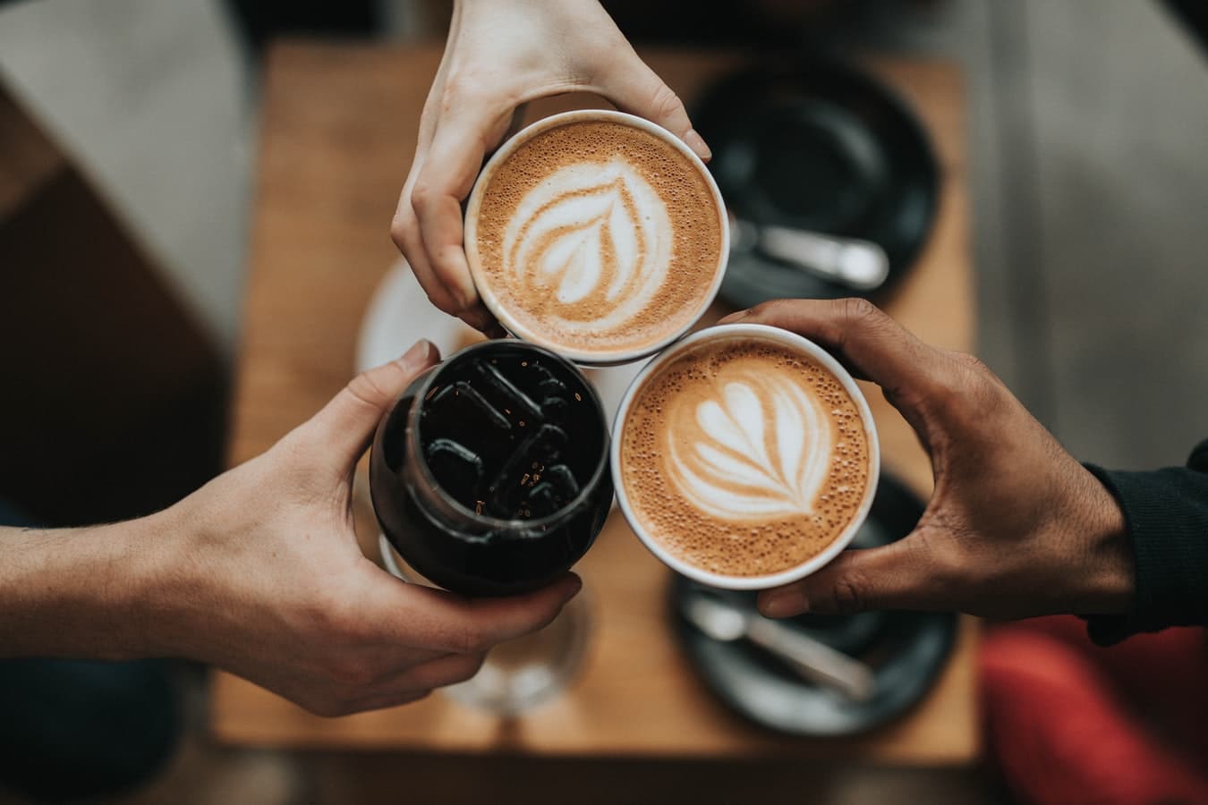 Three people toasting coffee drinks: two lattes with latte art and one iced coffee.