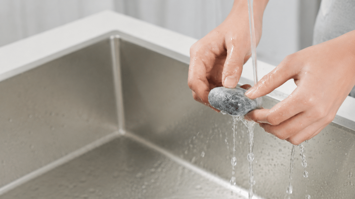 Hands washing a pumice stone under running water in a stainless steel sink.
