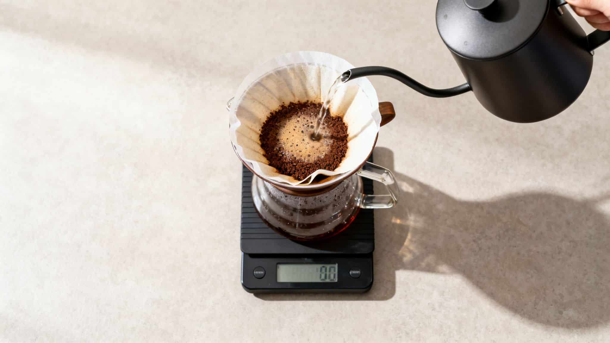 Overhead shot of pouring hot water from a black kettle into a coffee filter cone on a scale.