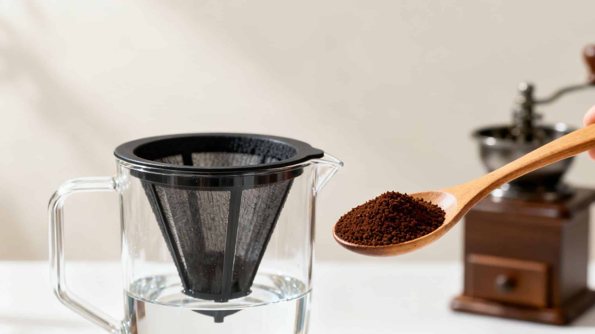 A hand holds a wooden spoon with coffee grounds over a dripper carafe with water.