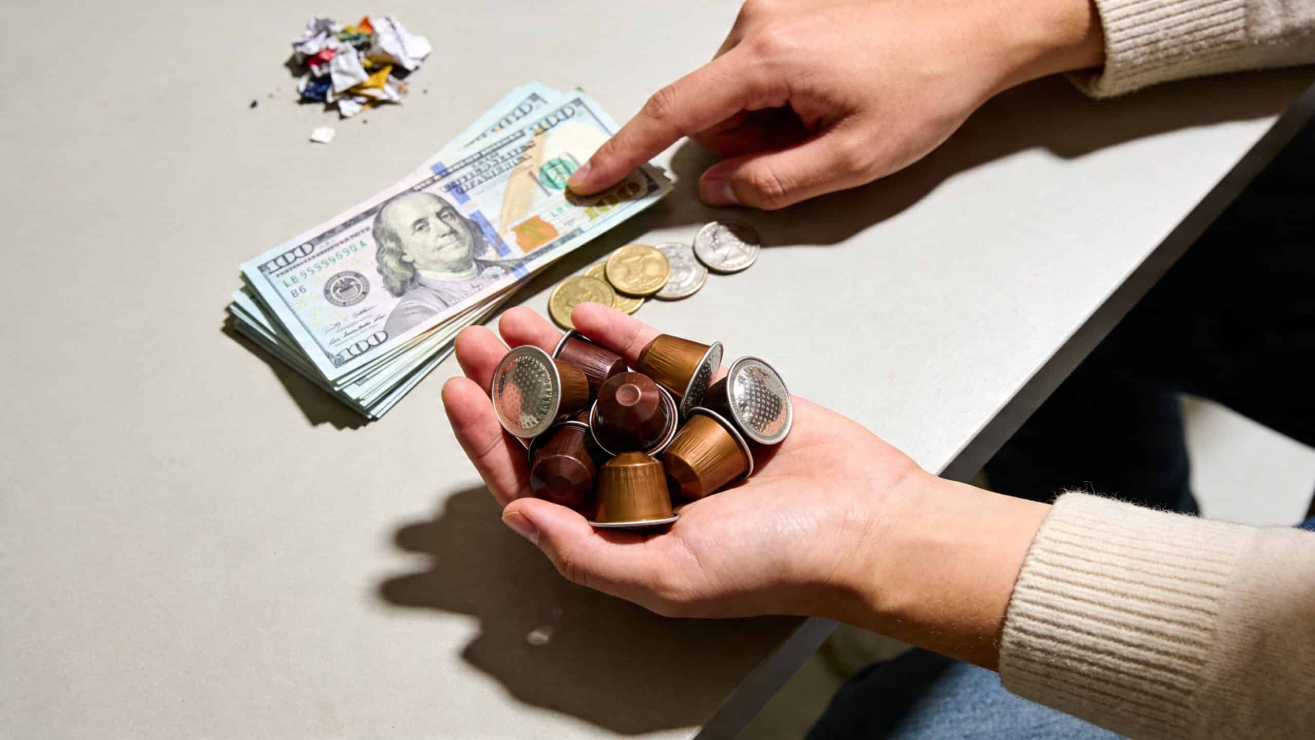 A person holds Nespresso coffee capsules in their hand next to a stack of American dollar bills.