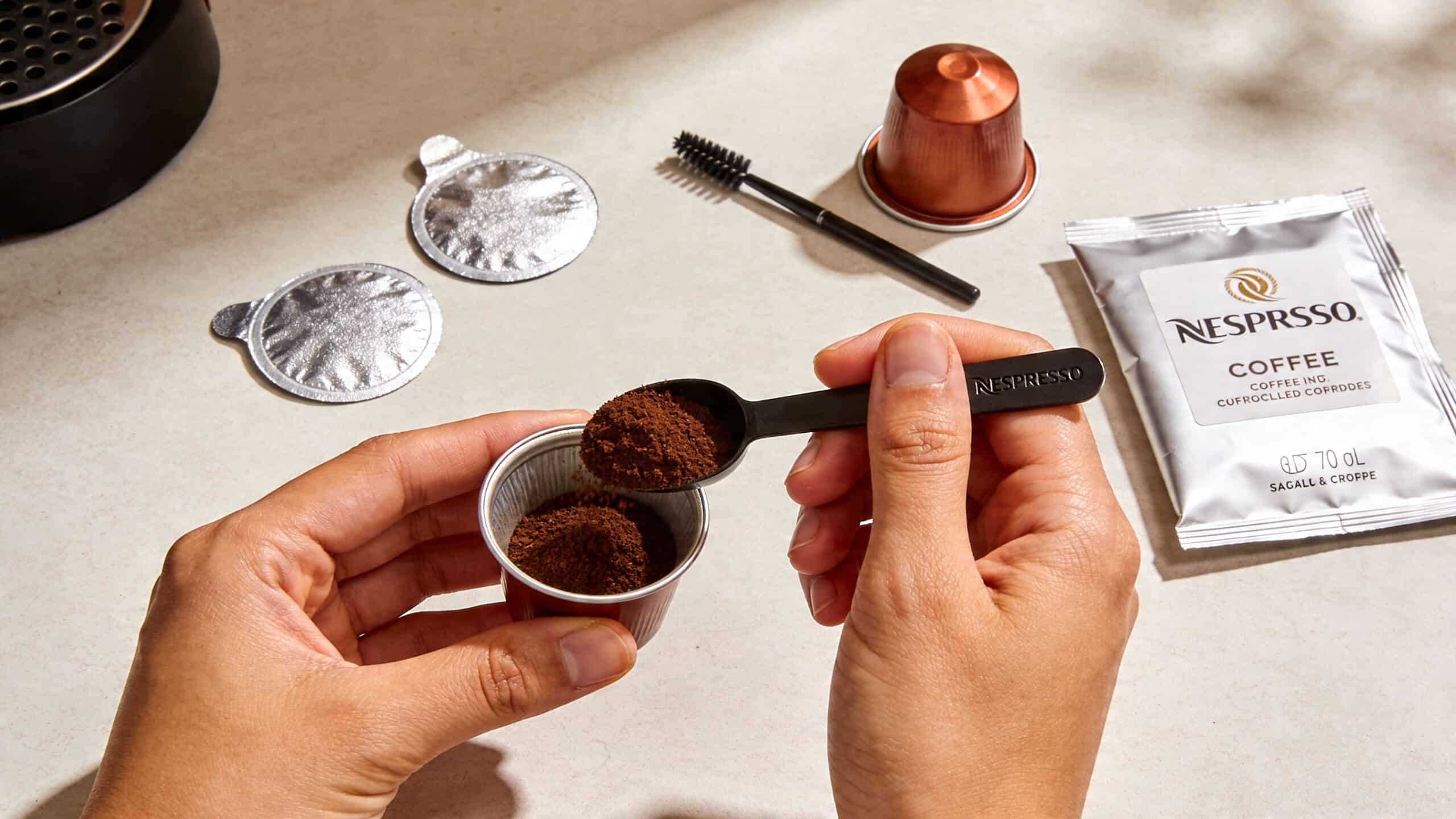 A person uses a Nespresso branded spoon to fill a reusable coffee capsule with ground coffee powder.
