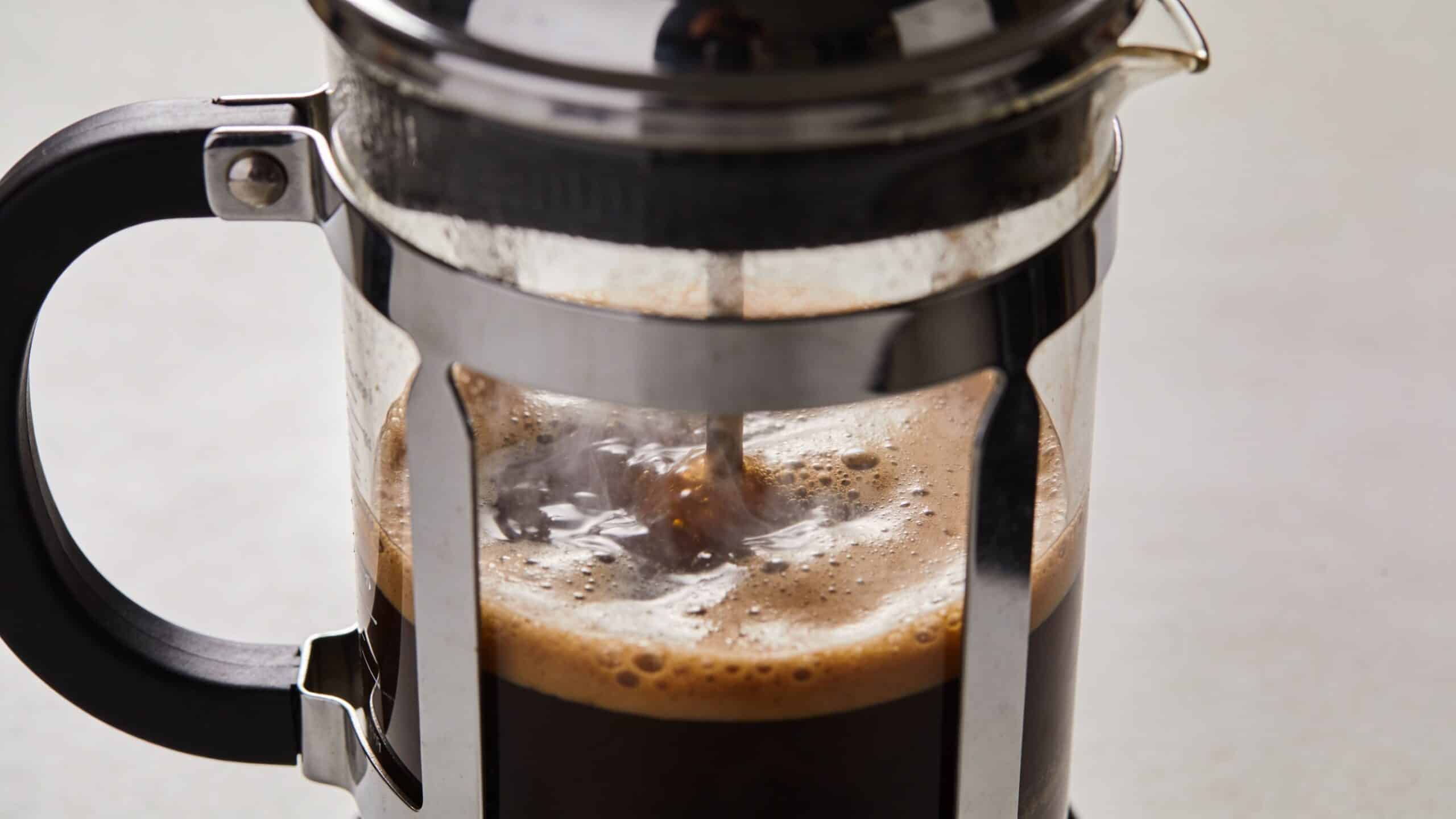 A close-up view of a metal French press brewing fresh coffee with visible steam and foam.