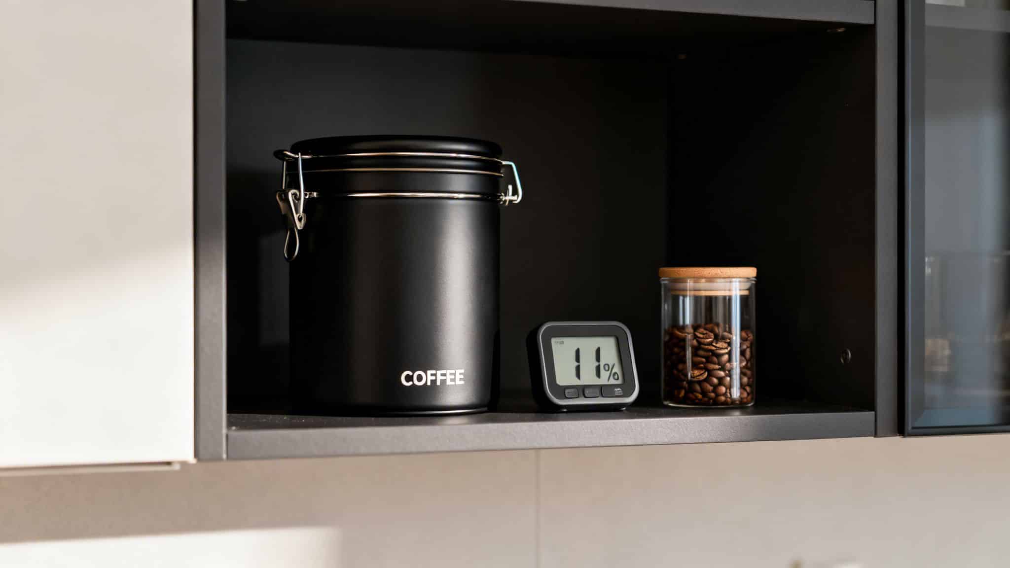 Black coffee container, digital hygrometer displaying 11%, and glass jar of coffee beans on a dark shelf.