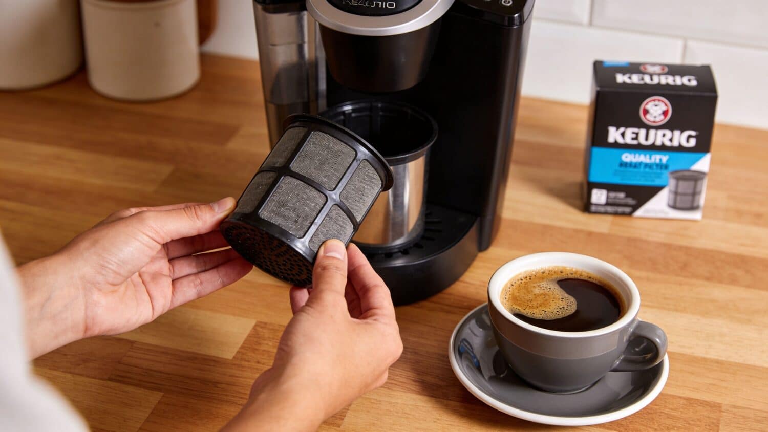 Person cleaning a reusable Keurig coffee filter next to a coffee maker and cup
