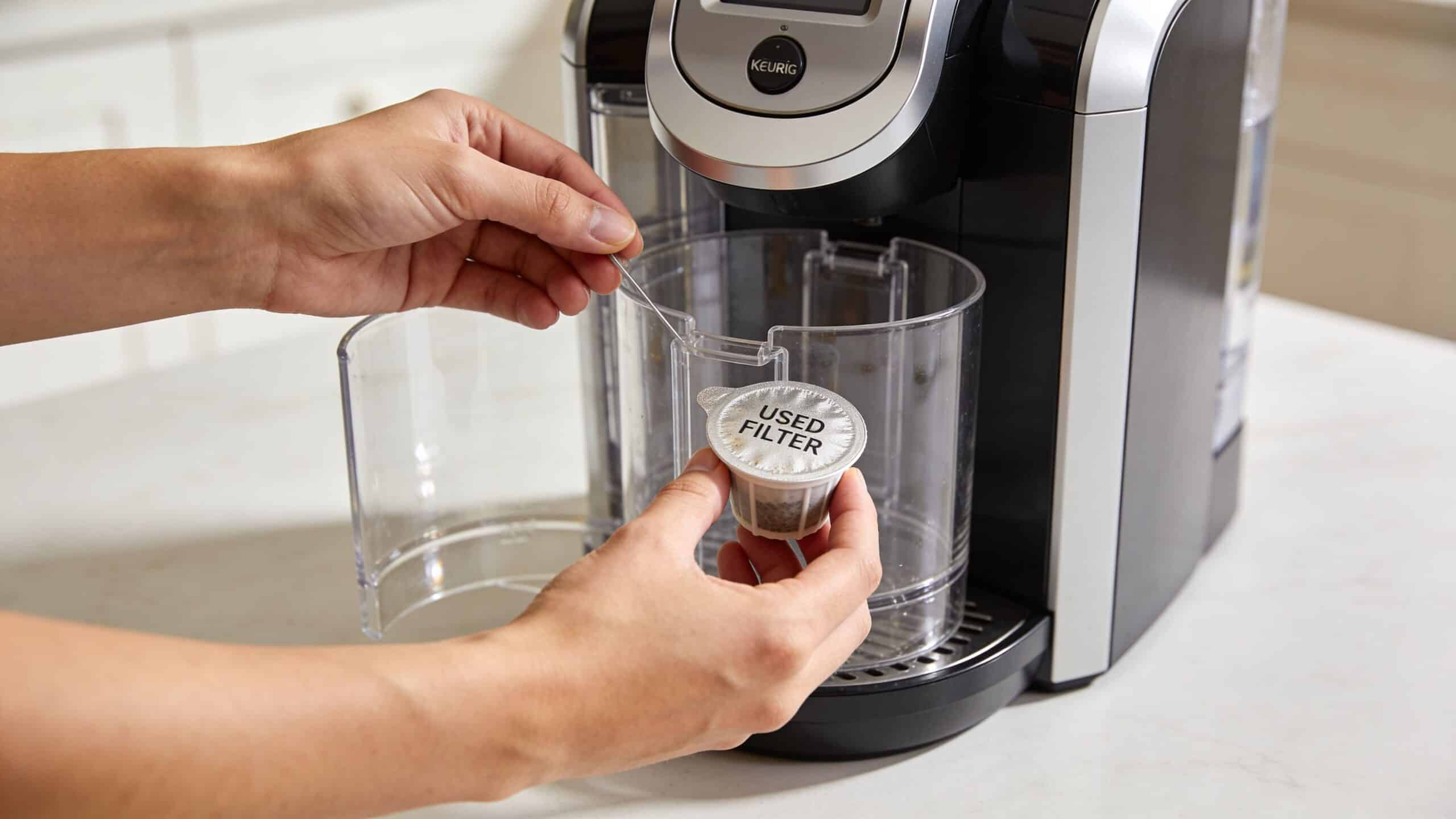 A person holding a used coffee filter pod in front of an open Keurig coffee machine.