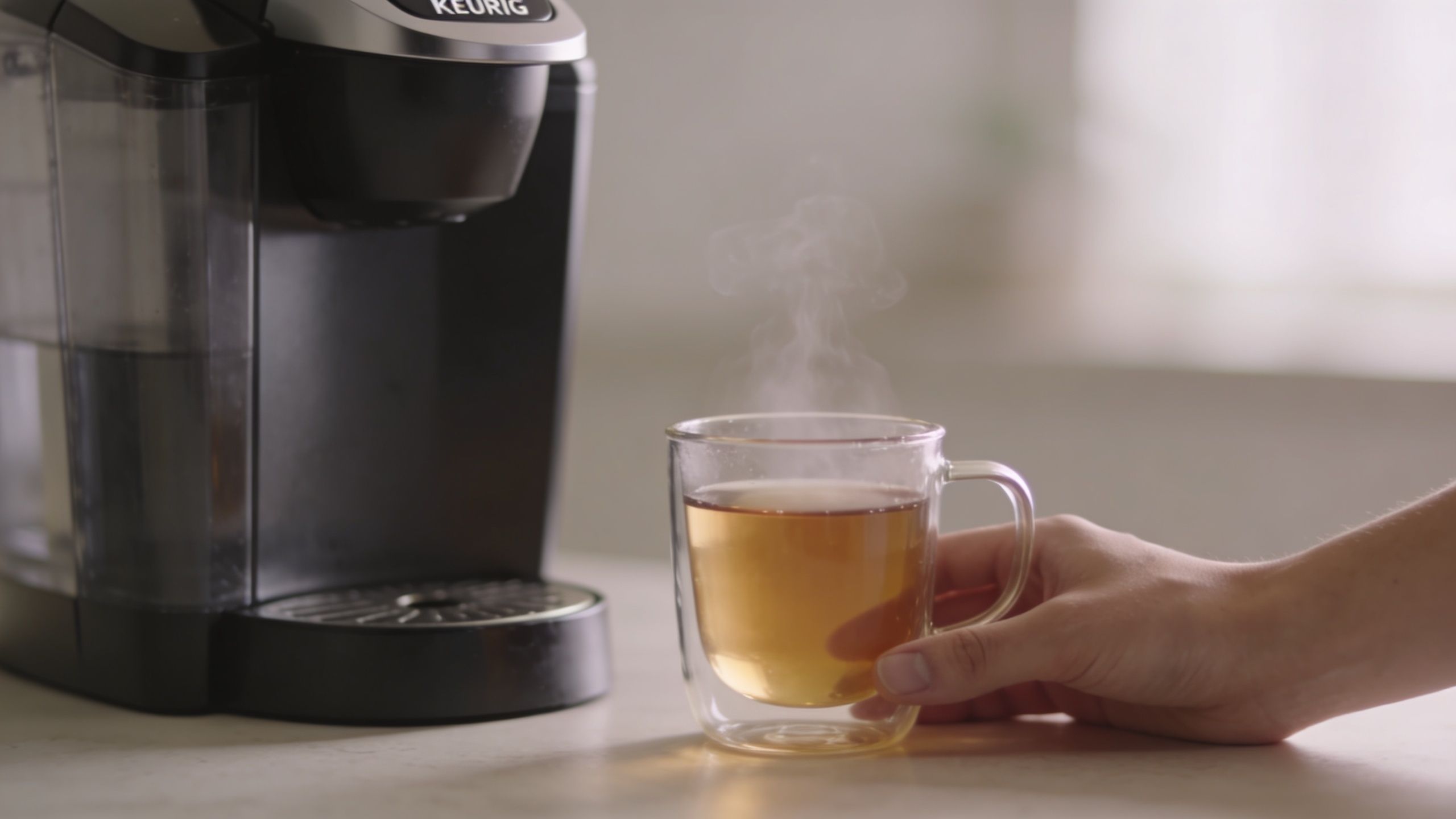 A hand holding a clear glass mug of hot tea beside a black Keurig coffee machine.