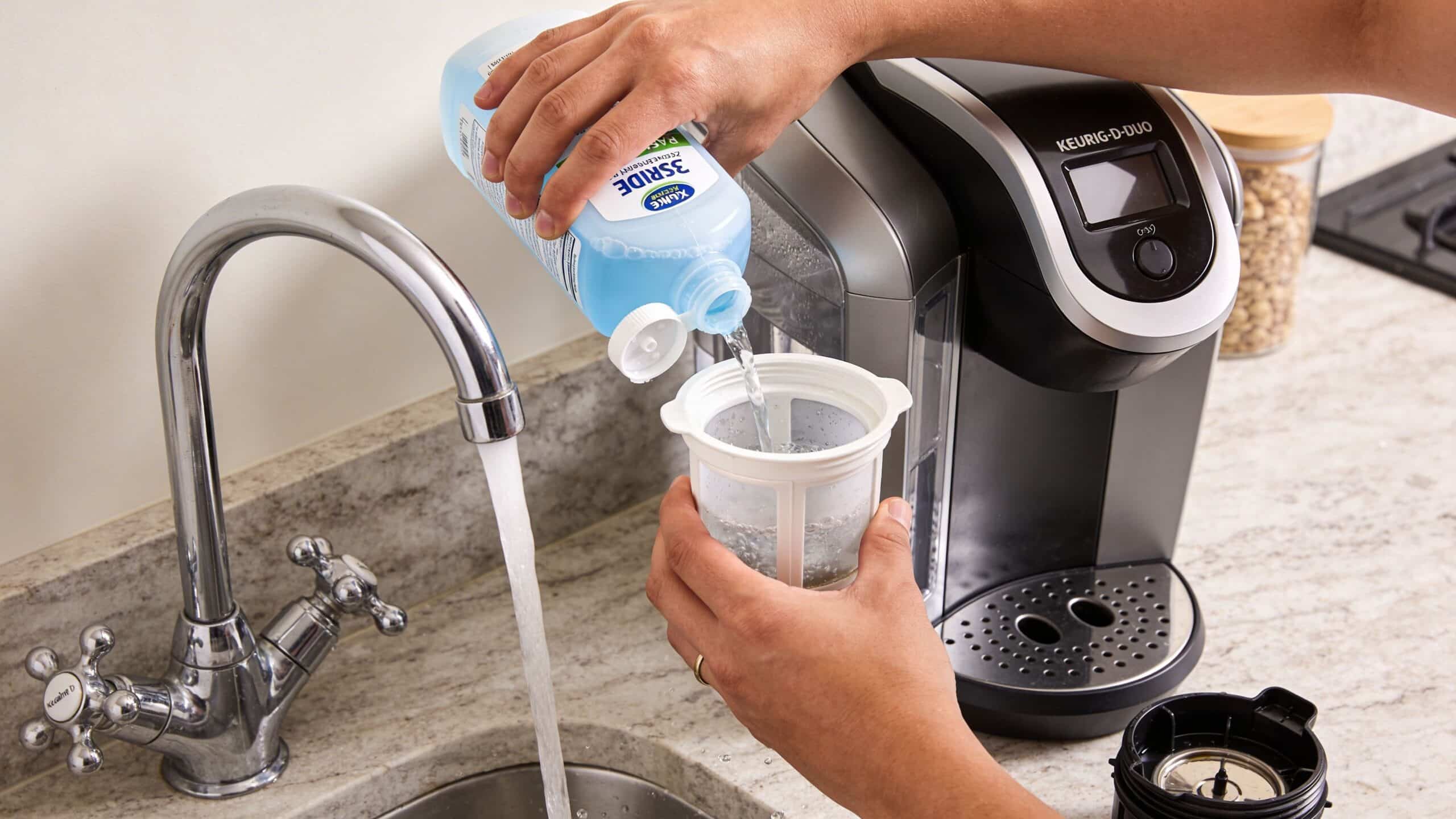 A person pouring cleaning solution from a blue bottle into a coffee machine reusable filter basket.