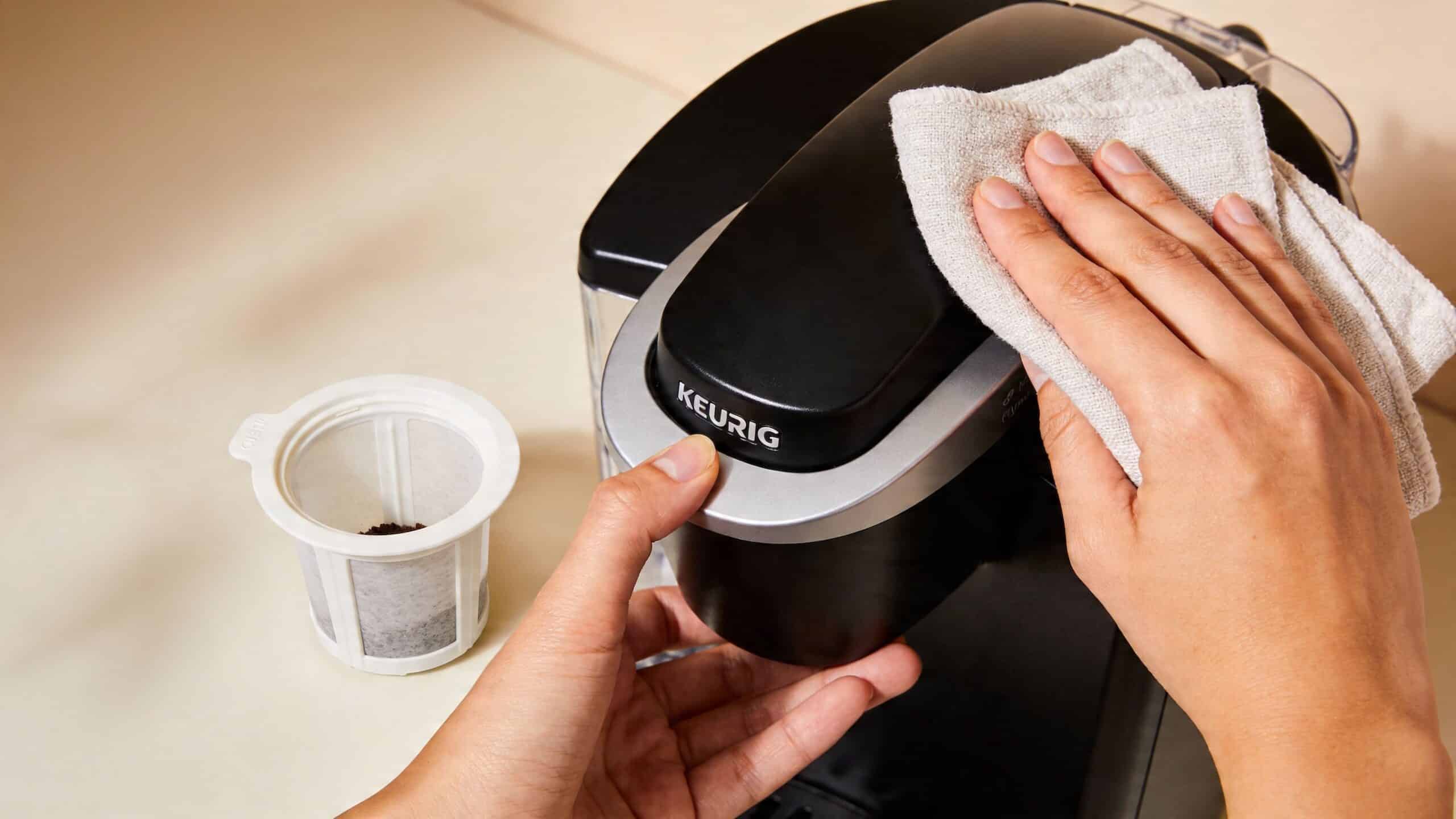 A person uses a beige microfiber cloth to clean a Keurig coffee maker while holding the lid.