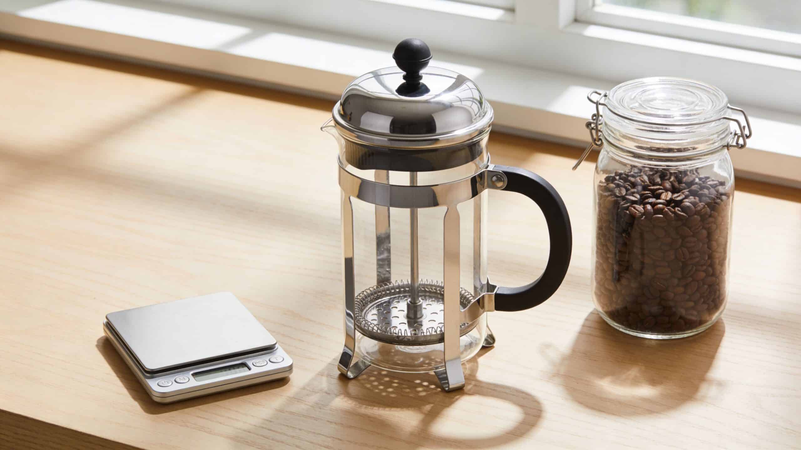 A stainless steel French press, a digital scale, and a jar of roasted coffee beans on wood.