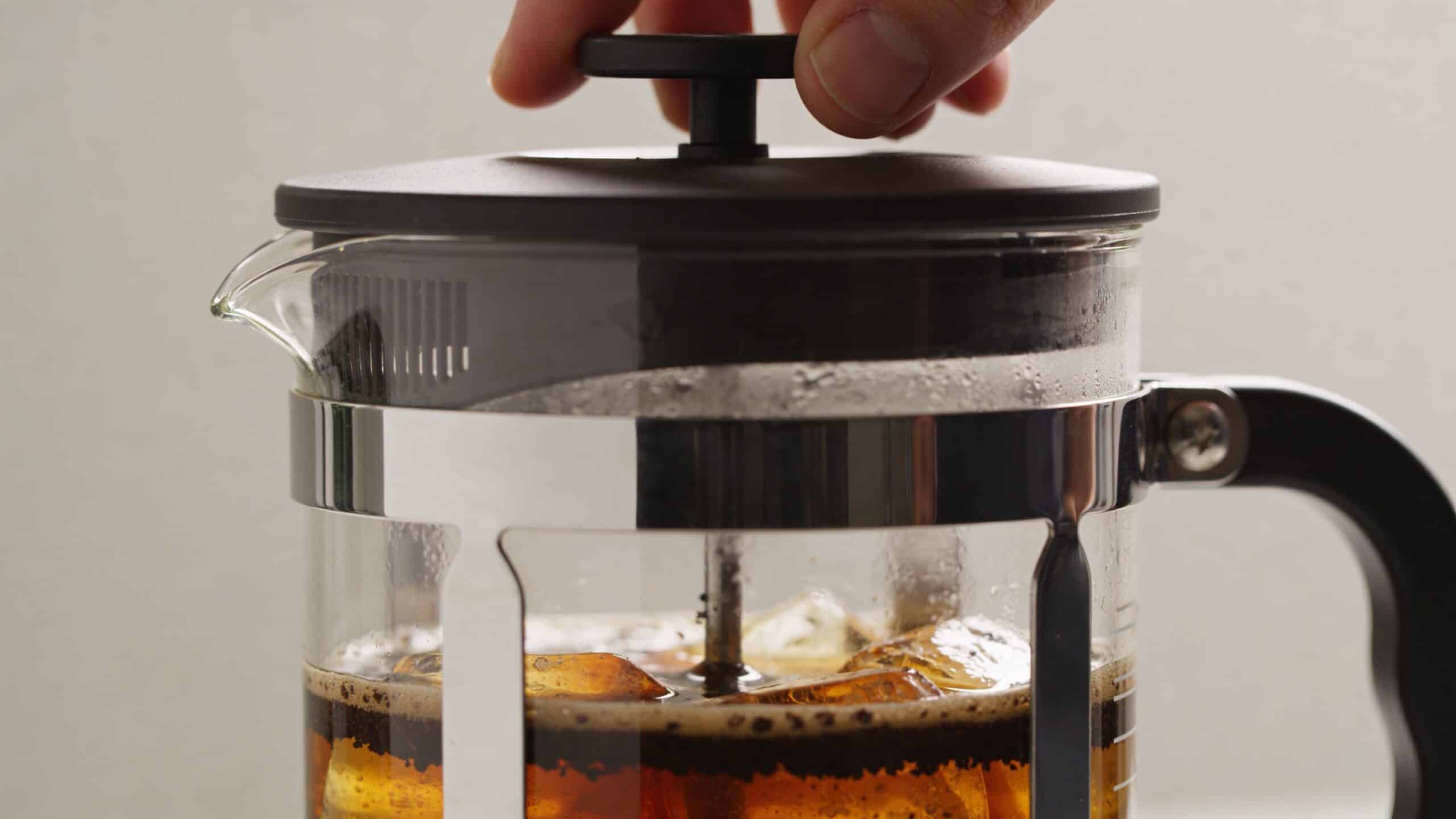 A hand pressing down the plunger of a French press filled with cold brew coffee and ice.