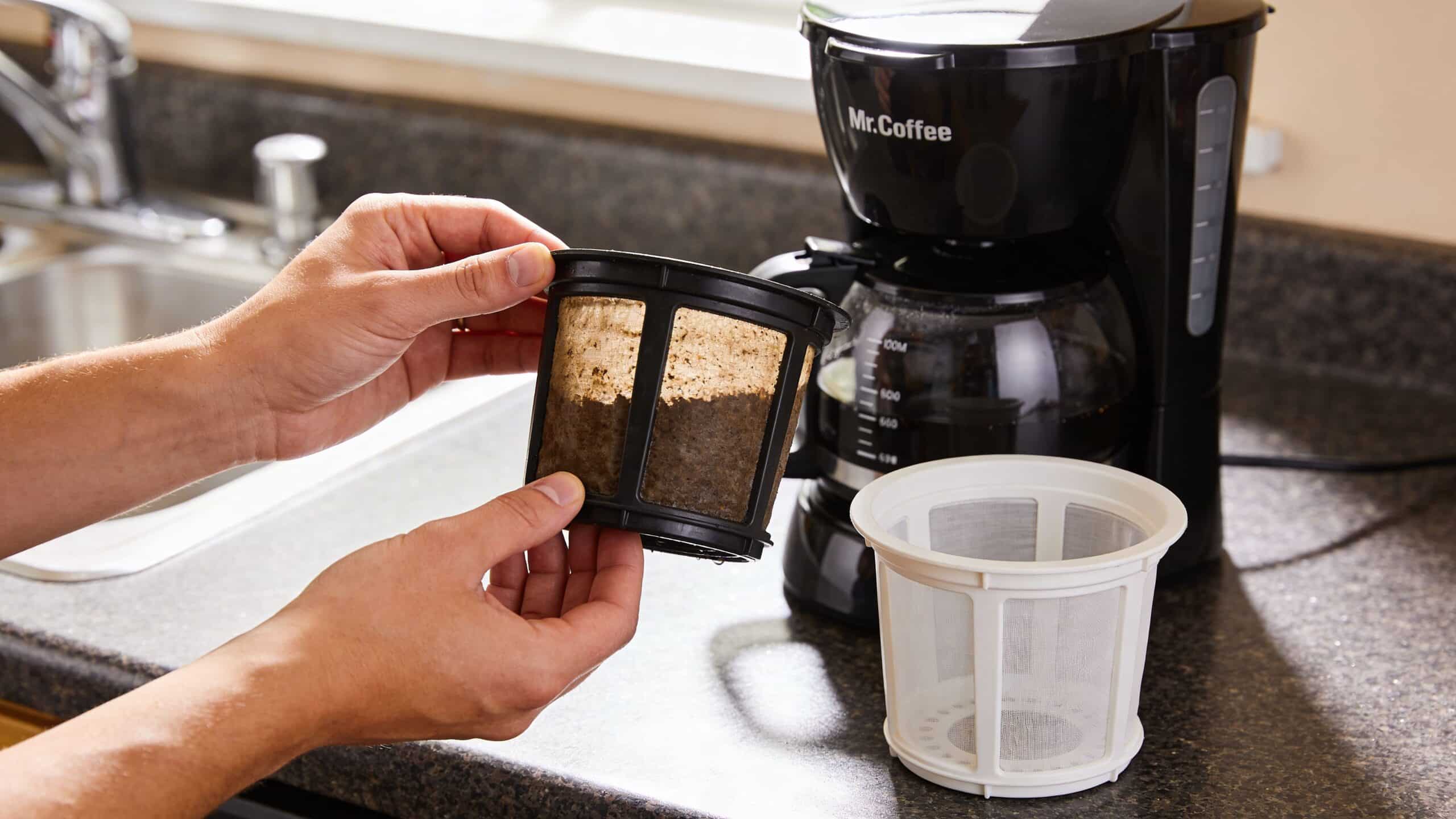 A person holds a used coffee filter next to a clean white filter and a coffee machine.