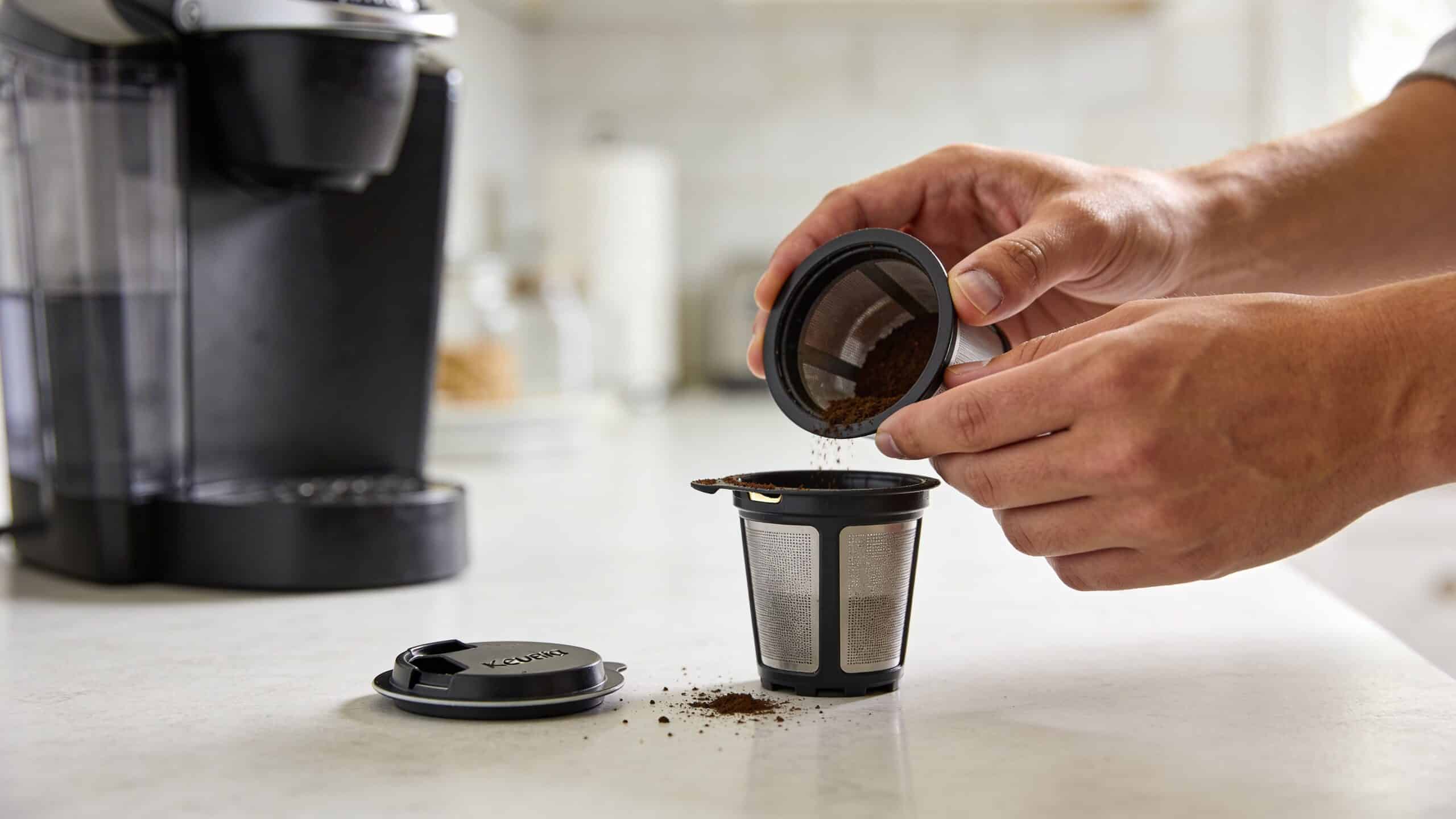 A person pouring coffee grounds into a refillable K-Cup filter basket on a kitchen counter.