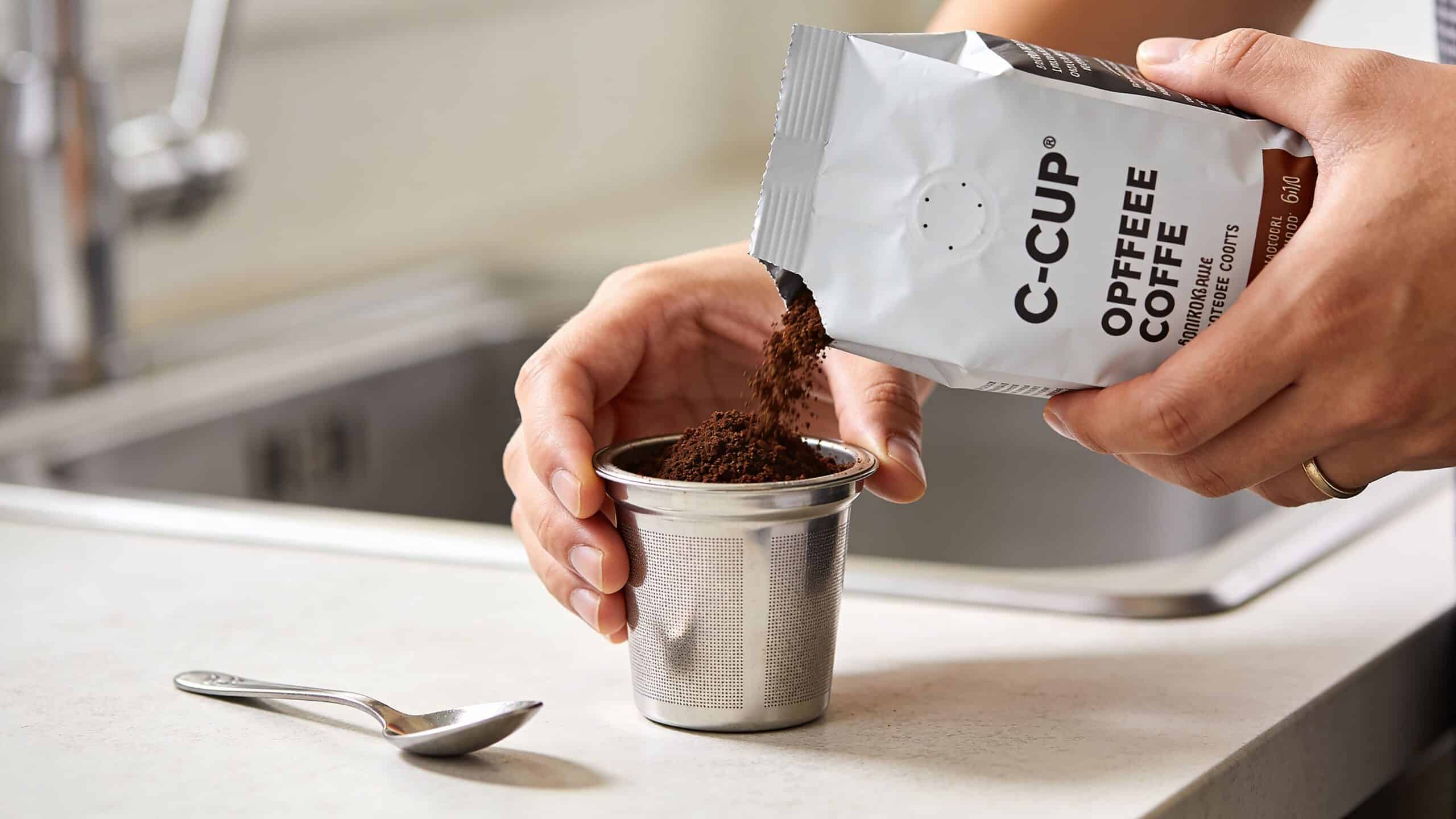 A person pouring ground coffee into a metal reusable coffee pod in a home kitchen setting.