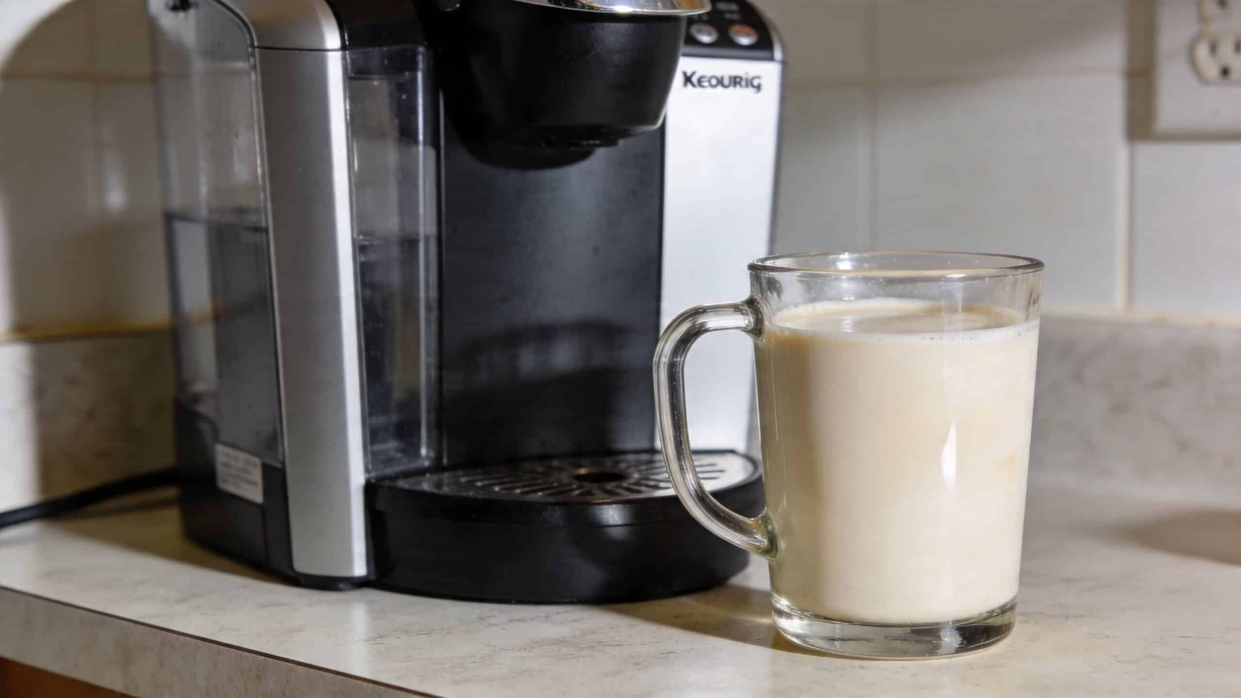 A glass mug filled with a creamy vanilla latte sitting beside a black Keurig coffee machine.