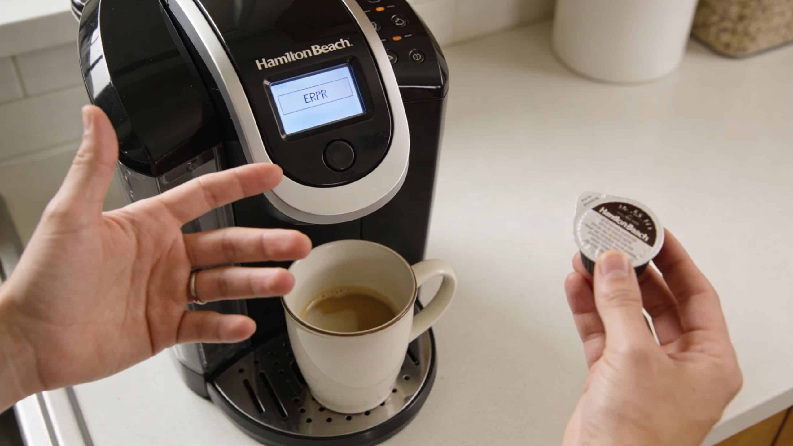 A person gesturing with frustration toward a Hamilton Beach coffee maker displaying an error code with pods.