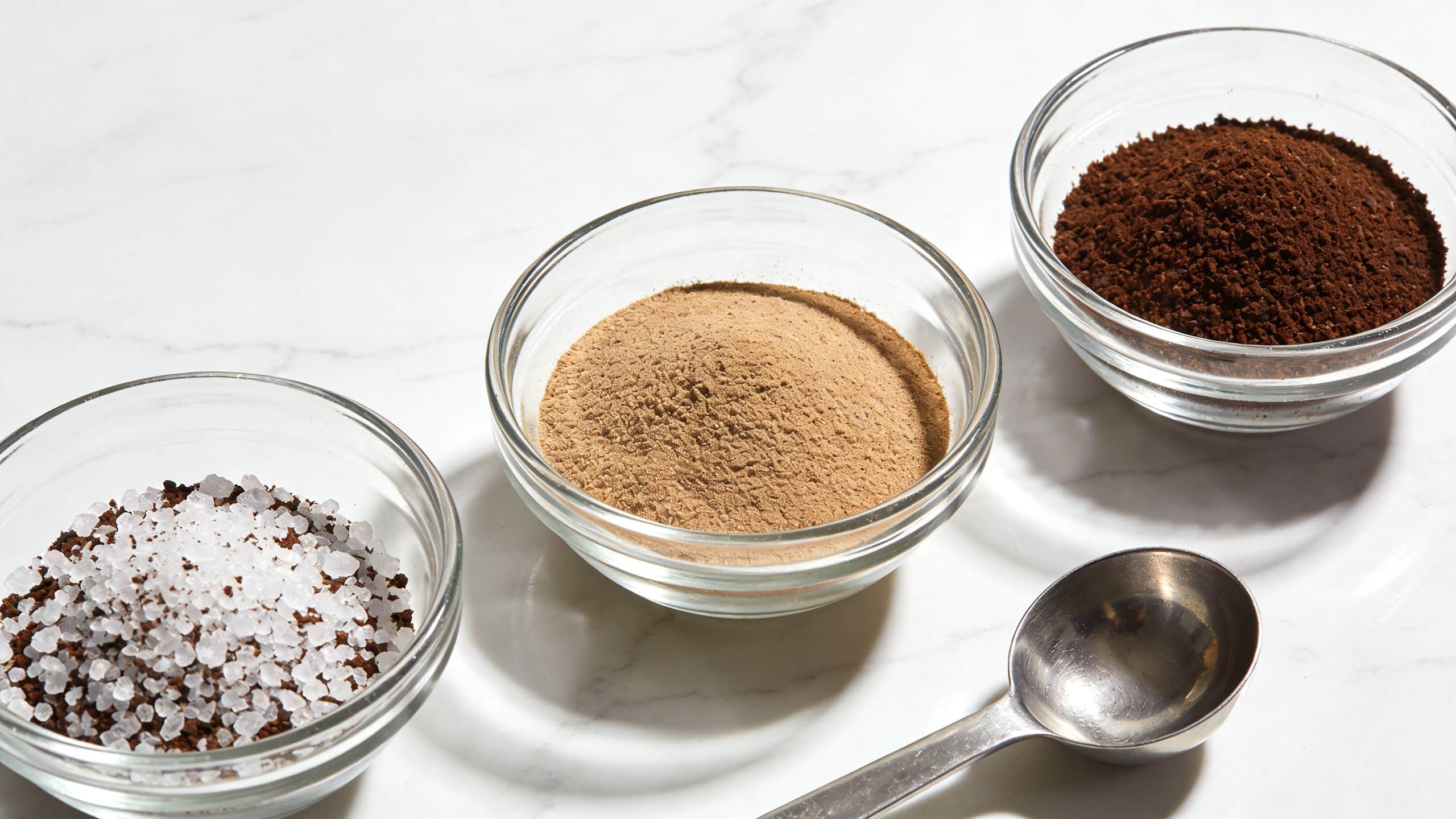 Three small glass bowls containing sea salt, ground coffee, and powdered spices arranged on a marble surface.