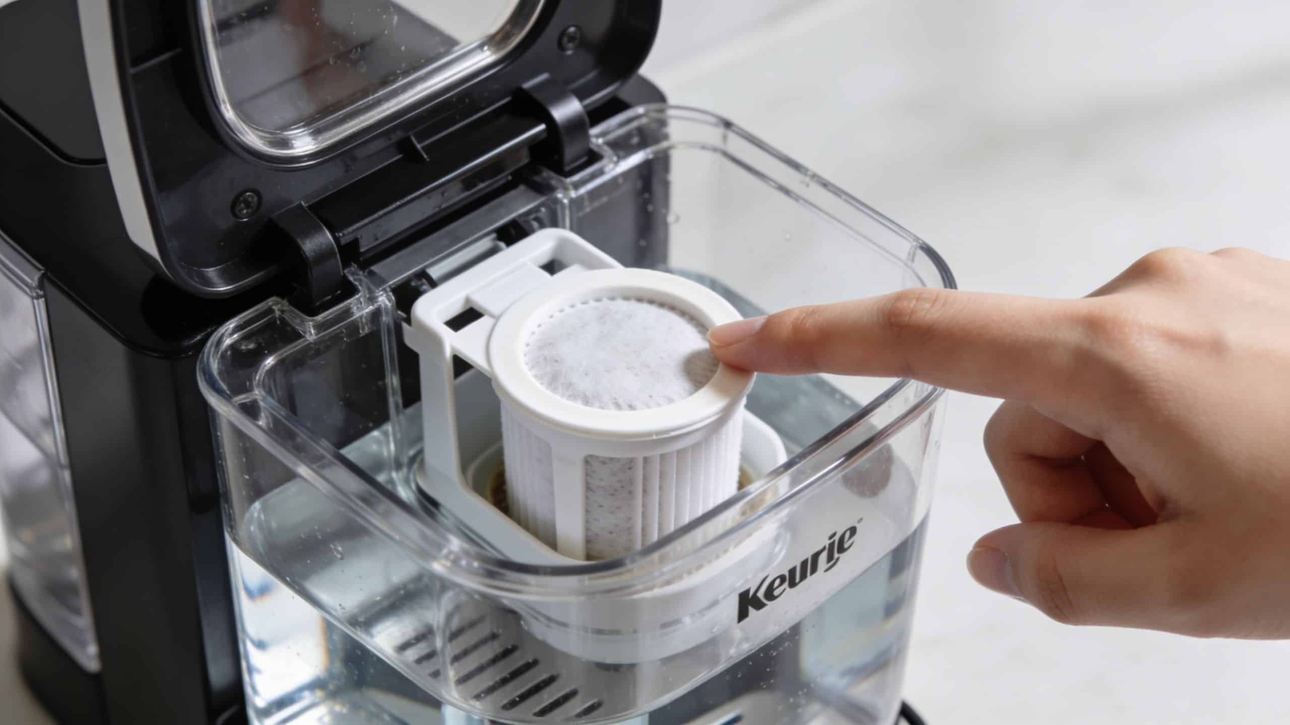 A person's finger pointing at the water filter inside the clear water reservoir of a Keurig coffee machine.