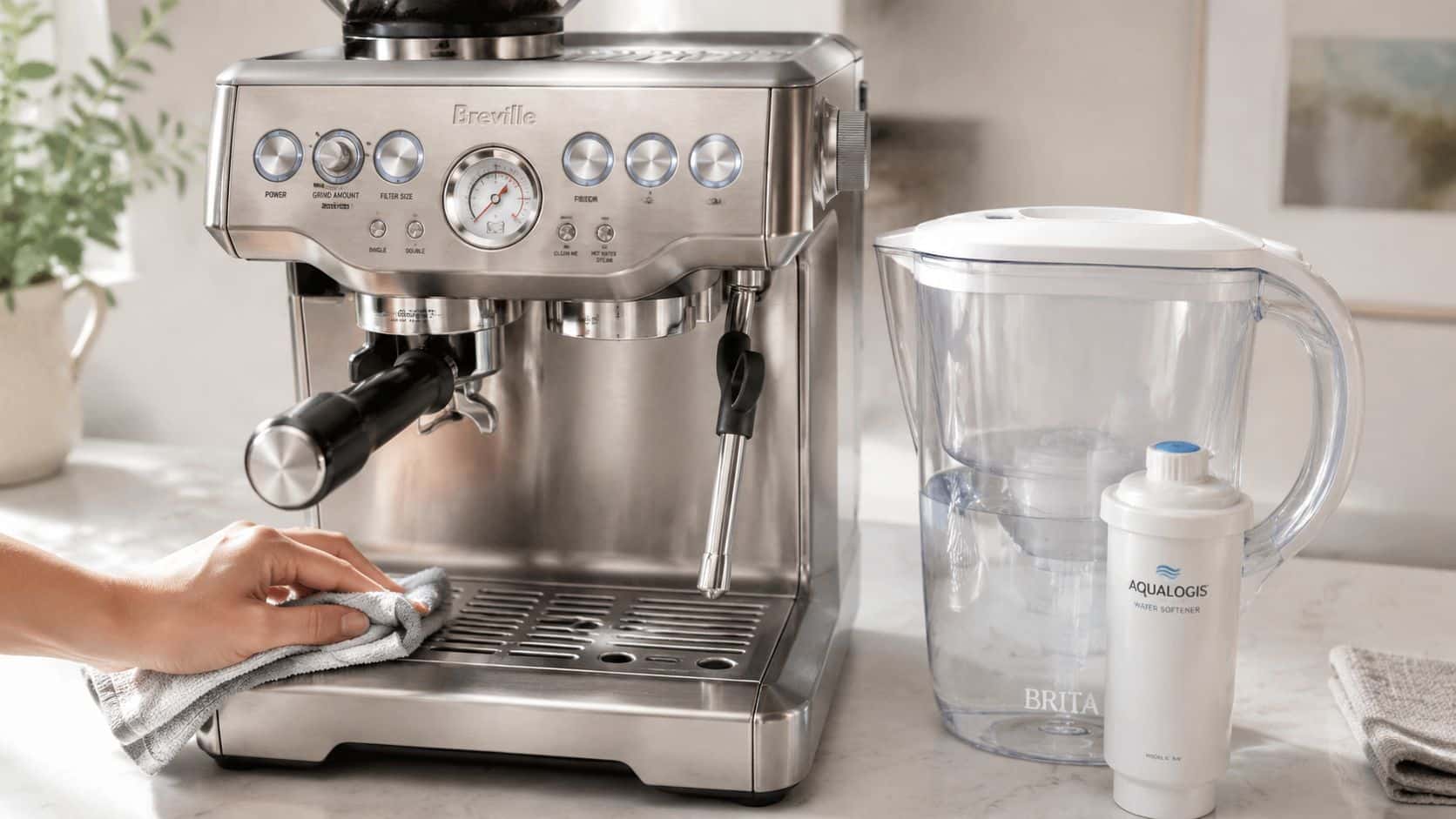 A hand wiping down a stainless steel Breville espresso machine next to a water filter pitcher.