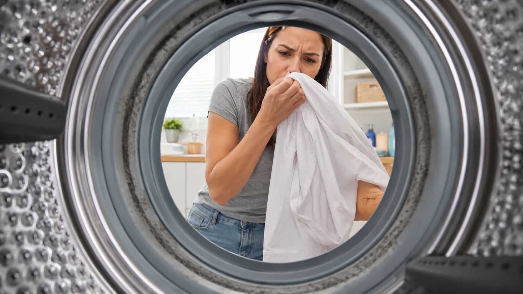 A woman looks concerned while smelling a piece of white clothing taken from inside a washing machine.