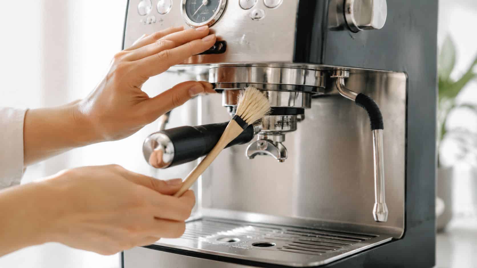 A close-up view of a person using a small wooden brush to clean an espresso machine's group head.
