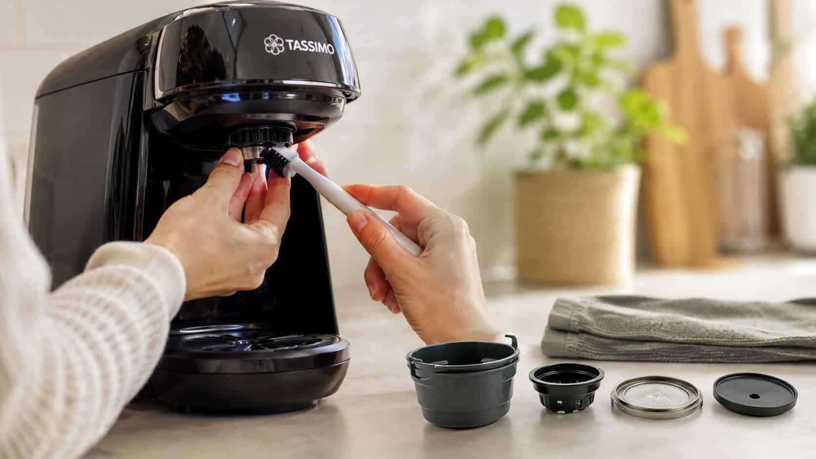 A person cleaning the Tassimo coffee machine brew head with a small brush alongside reusable coffee pods.