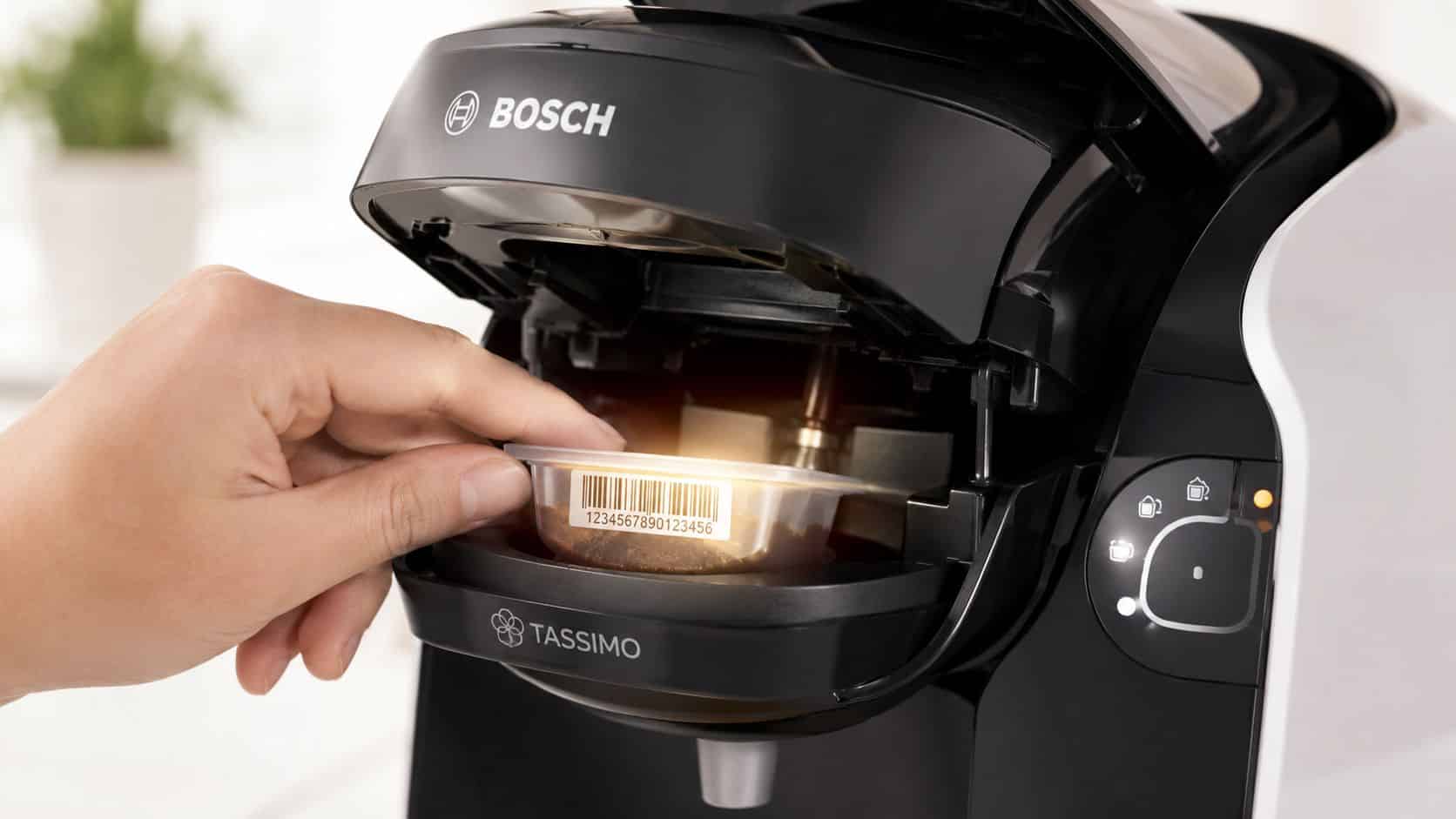 A person placing a Tassimo compatible refillable coffee pod into a black Bosch coffee machine for brewing.