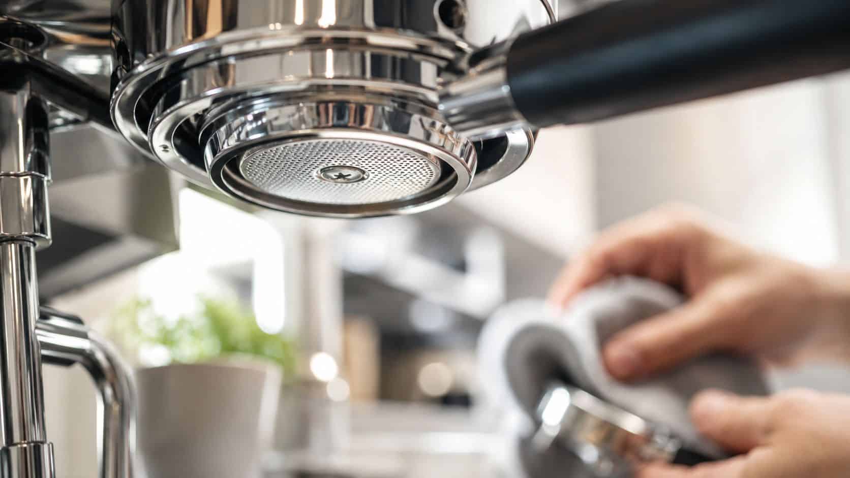 A close-up view of an espresso machine group head while a person cleans a portafilter.