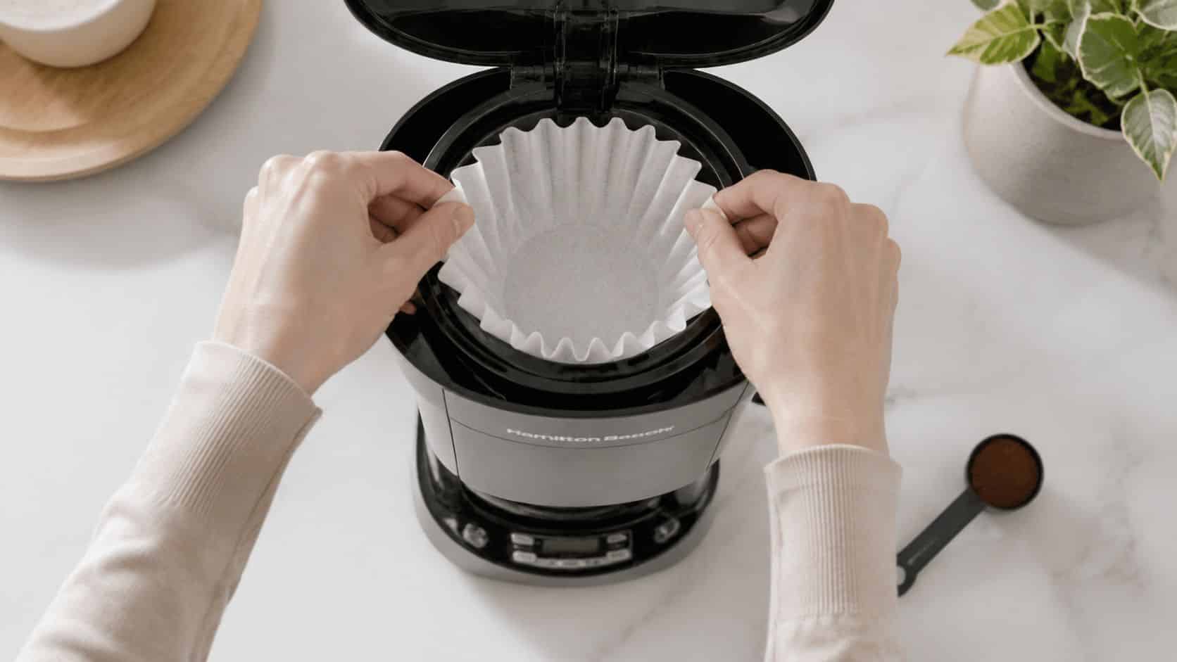 A person placing a paper coffee filter into a Hamilton Beach coffee maker on a marble countertop.