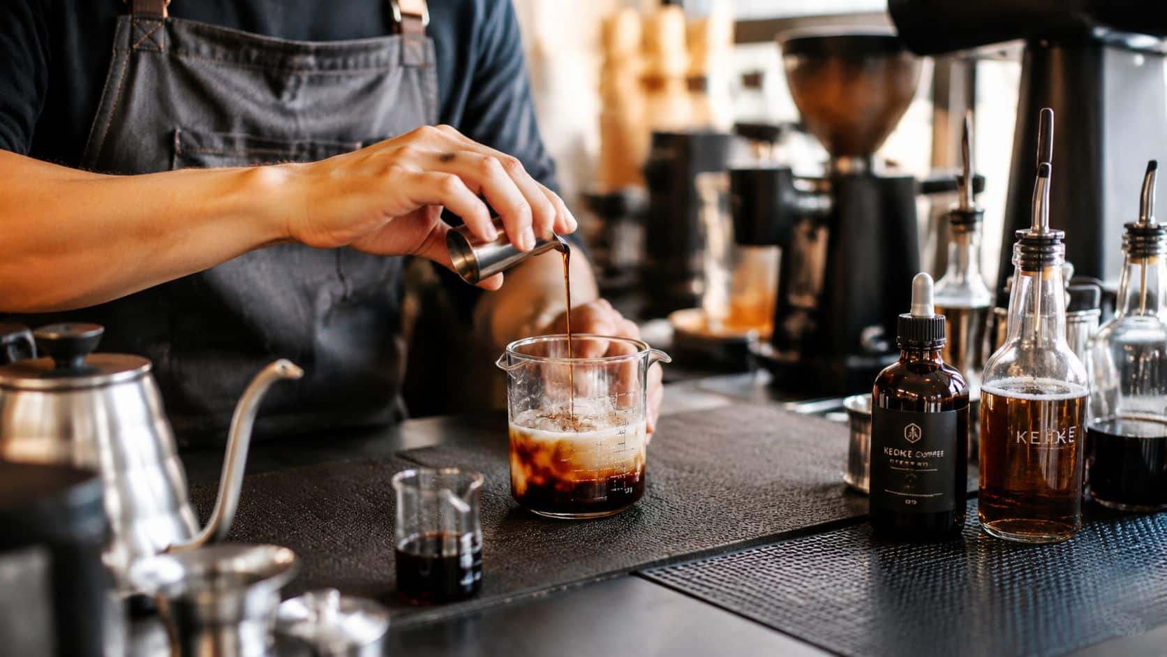A barista pouring a shot of concentrated coffee into a measuring glass filled with ice and milk.