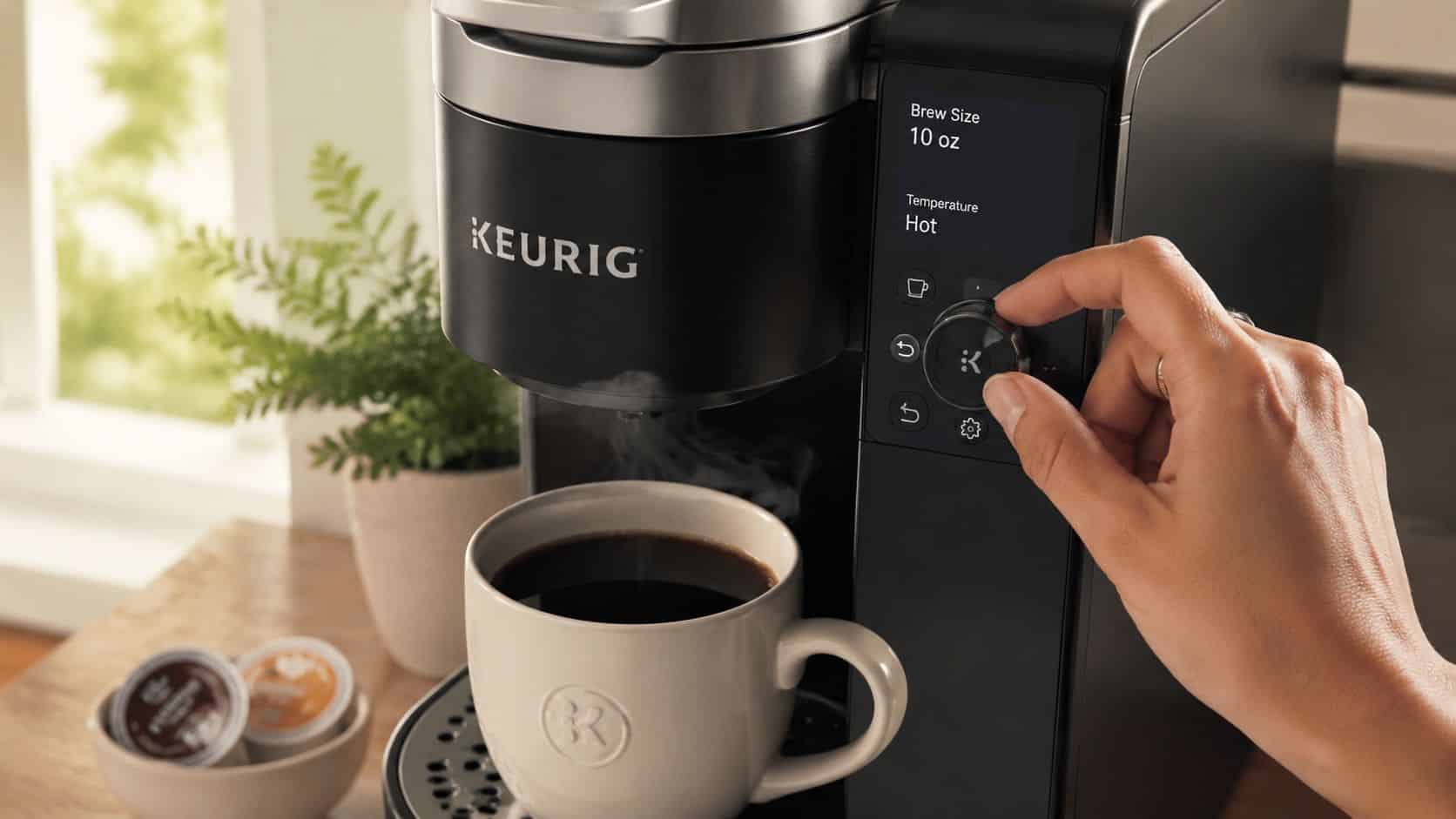 A hand adjusts the dial on a black Keurig coffee maker brewing a cup of hot coffee.