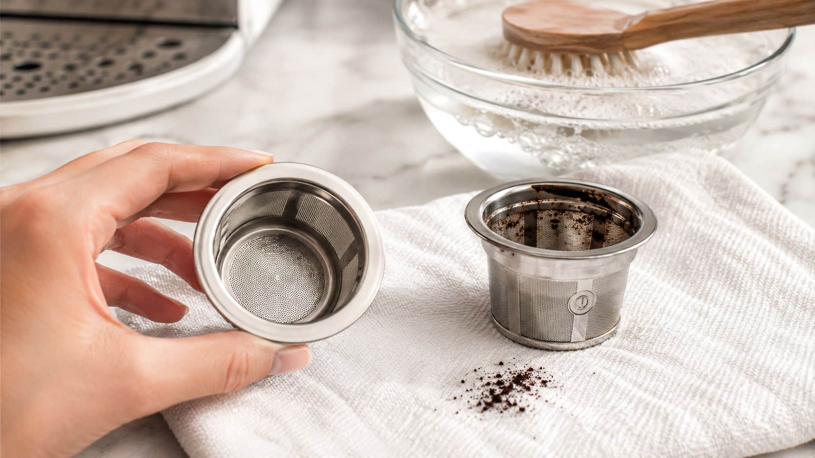 A person holding a clean stainless steel coffee capsule filter next to a used one on a towel.