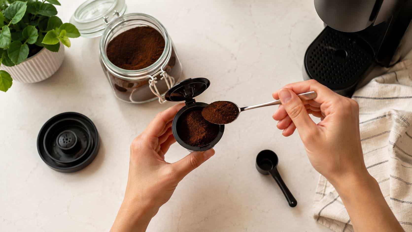 A person fills a reusable coffee pod with ground coffee using a small spoon on a table.
