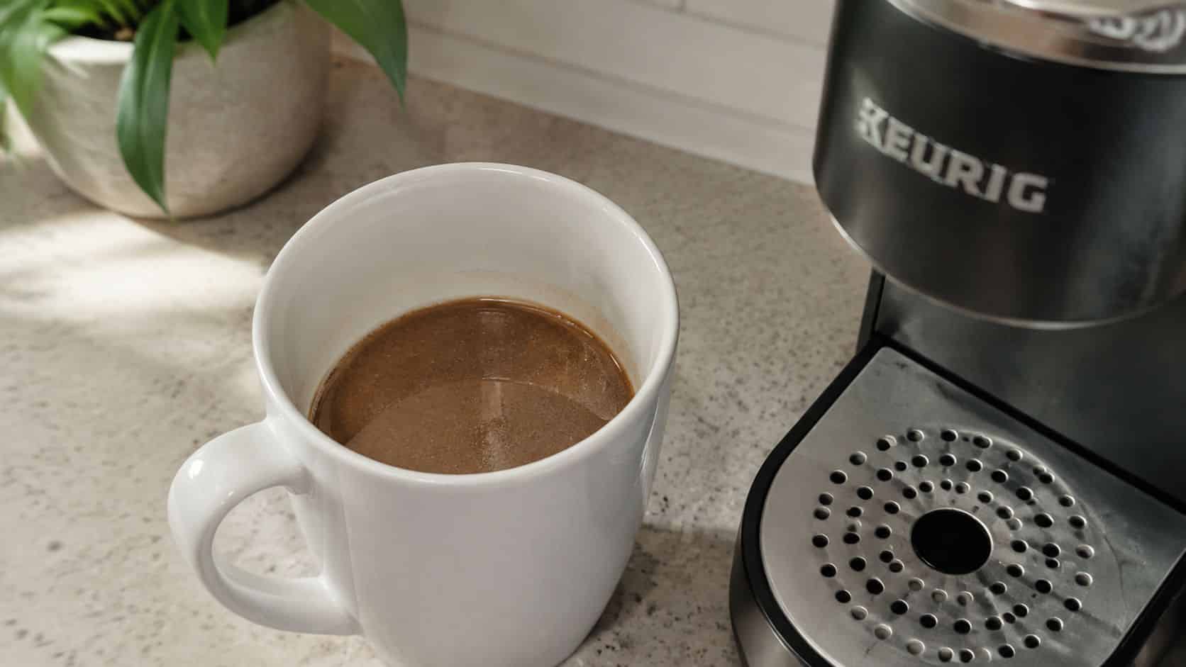 A white ceramic mug filled with fresh coffee next to a black Keurig coffee machine on a counter.