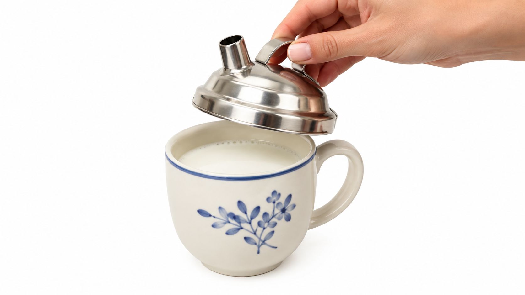 A hand placing a stainless steel cover with a small spout onto a decorative porcelain milk mug.
