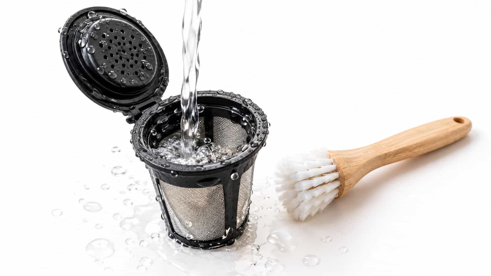 A reusable coffee pod being rinsed with water under a faucet next to a wooden cleaning brush.