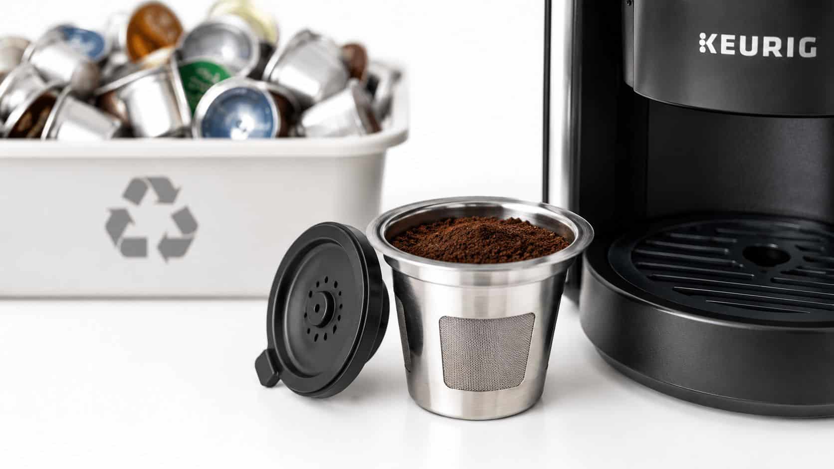 A stainless steel reusable coffee pod filled with coffee grounds next to a Keurig machine and bin.