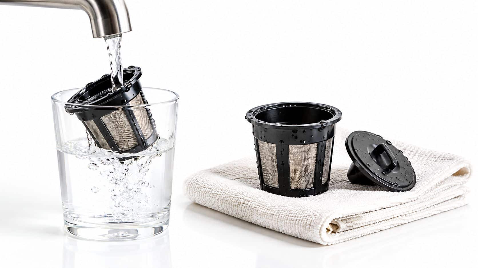 A reusable coffee pod filter being rinsed under a faucet into a glass of water on a towel.