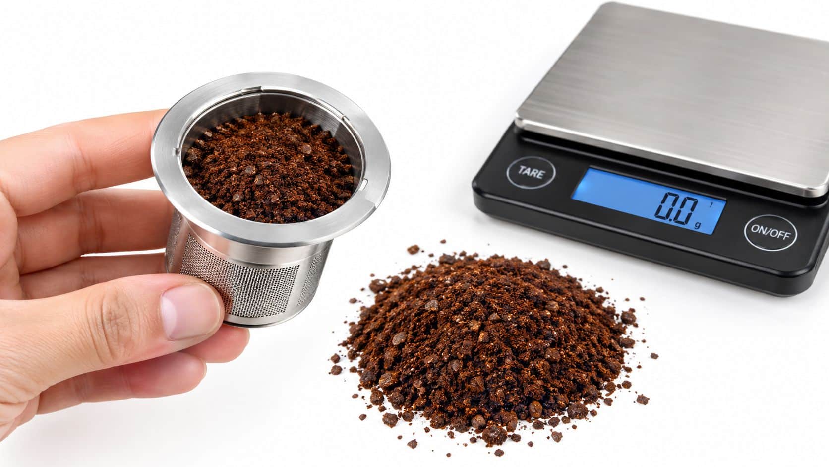 A hand holding a stainless steel coffee pod filled with ground coffee next to a digital scale.